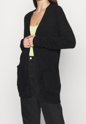 Femme portant un cardigan noir duveteux ouvert sur un haut vert clair et un jean noir taille haute, les mains jointes près de la poitrine.