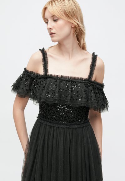 Robe noire avec un corsage en sequins ajusté, des volants en tulle superposés au niveau du décolleté et une jupe fluide au fini texturé. Les bretelles sont effilochées.