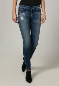Jeans ajustados de mezclilla azul oscuro, con detalles desgastados y adornos cerca de los bolsillos. Talle medio con una silueta entallada.