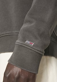 Šedá mikina vyrobená z měkkého materiálu s žebrovanými manžetami. Obsahuje malé vyšívané logo v červené, bílé a modré barvě na rukávu.