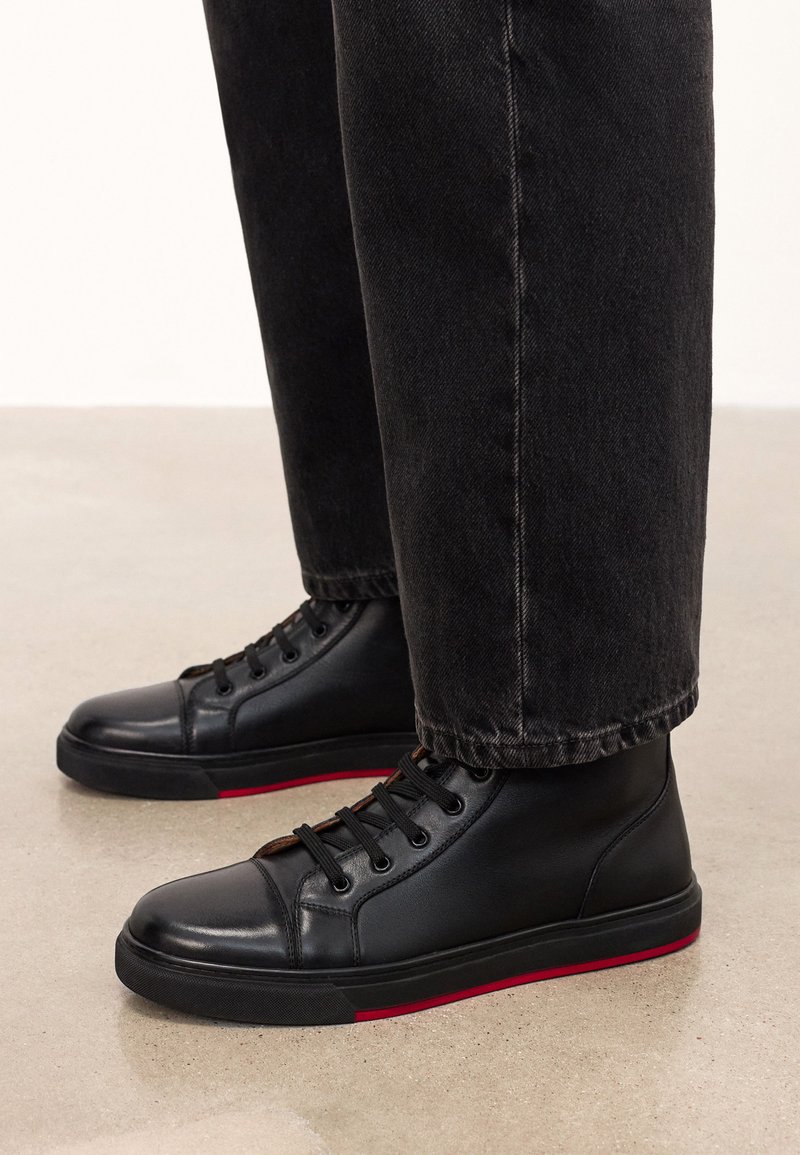 Mustad musta nahast kõrge varrega tossud punaste aktsentidega, ümmarguste varvastega, seotud kingade disainiga ja tekstuuriga kummist tallaga, sobitatud tumedate teksadega.