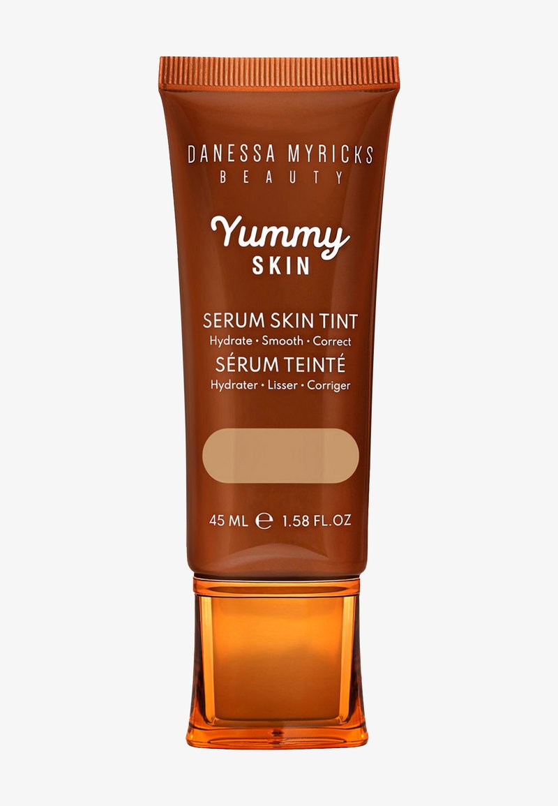 Tube marron avec un capuchon brillant, étiqueté "Yummy Skin Serum Skin Tint." Contient 45 mL. Texture lisse et couvrance moyenne indiquées.