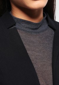 Veste noire à col structuré associée à un col roulé gris doux. Le col roulé présente une texture subtile, superposée sous la veste.