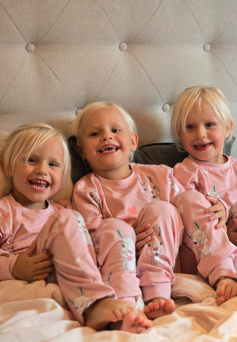 Three children wearing matching soft pink pajamas with bunny illustrations, sitting on a bed with a tufted headboard.