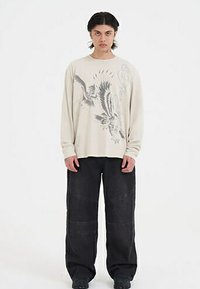 Langärmliges cremefarbenes Hemd mit Adler-Graphic, kombiniert mit locker sitzenden schwarzen Jeanshosen mit sichtbaren Nähten und großen Taschen.