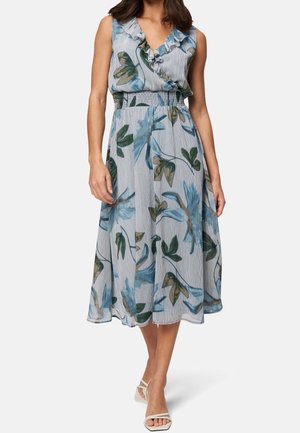 Femme portant une robe midi bleu clair sans manches avec un col à volants et un grand imprimé floral, associée à des talons blancs ouverts à l'avant.
