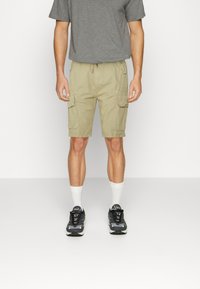 Beige cargo shorts dengan saku samping, waist dengan tali serut, dan potongan longgar, dipadukan dengan kaos abu-abu dan sepatu sneakers hitam dengan aksen putih.