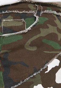 Tissu à motif camouflage dans des nuances de marron, de vert et de noir avec des bords effilochés. Comprend une section à motif contrastant blanc.