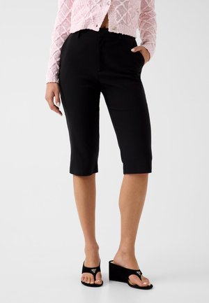 Mujer con pantalones cortos negros ajustados hasta la rodilla, un cárdigan rosa texturizado y sandalias negras de cuña con una trabilla para el dedo del pie.