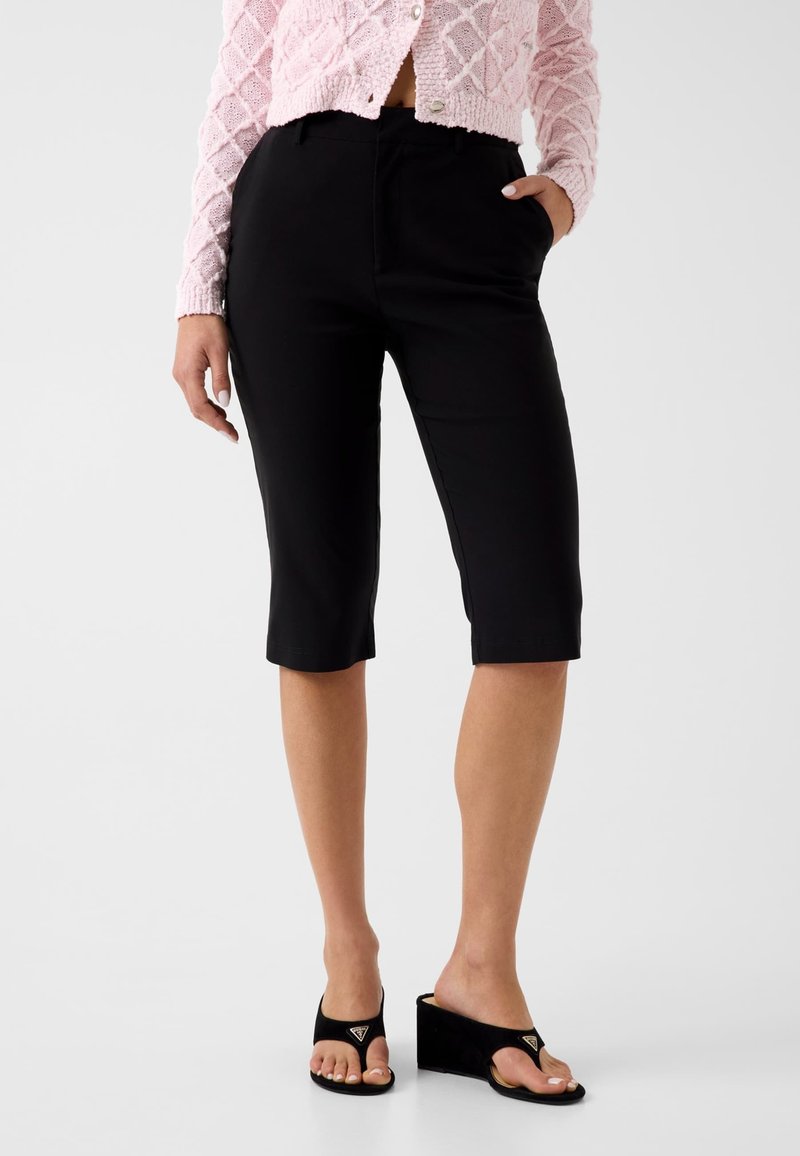 Femme portant un short noir ajusté jusqu'aux genoux, un cardigan rose texturé et des sandales compensées noires avec une bride pour l'orteil.
