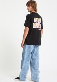 Camiseta gráfica negra con diseños de dibujos animados en rosa y amarillo en la parte posterior. Combinada con vaqueros anchos de denim azul desgastado y zapatillas negras.