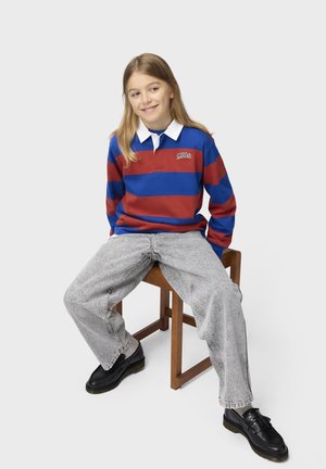 Persona joven sentada en un taburete de madera, vistiendo una camiseta polo con rayas azules y rojas, vaqueros gris claro y zapatos negros, sonriendo ligeramente.