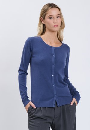Donna che indossa un cardigan blu attillato con bottoni e maniche lunghe e pantaloni scuri plissettati, posa con le mani nelle tasche davanti a uno sfondo semplice.