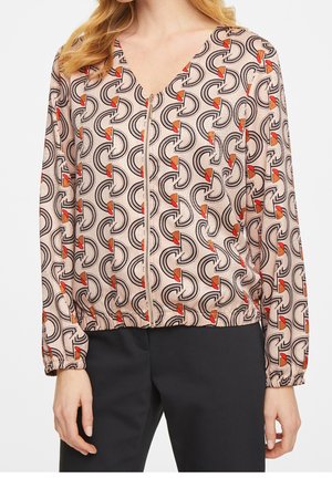 Kvinde iført en beige bluse med lynlås og sorte og orange geometriske mønstre samt sorte bukser, stående foran en hvid baggrund.