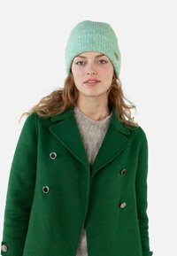 Miętowo zielona dziergana czapka o prążkowanej teksturze, noszona z zielonym dwurzędowym płaszczem i szarym swetrem. Okrągłe srebrne guziki na płaszczu.