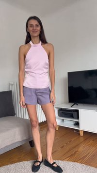Vaaleanpunainen satiininen halterneck-toppi, yhdistetty laventelinvioletteihin shortseihin. Mustat kengät, seisoo puulattialla, taustalla vaalea matto ja huonekaluja.