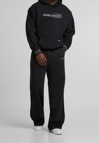Černá mikina s kapucí a tepláky vyrobené z měkké látky, s velkým logem na hrudi a jemnými pruhovanými akcenty na manžetách. Bíl�é tenisky.
