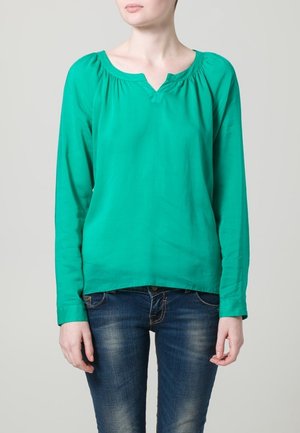 Groene longsleeve top met V-hals, voorzien van geplooide details bij de schouders en een gladde, lichte stof. Gepaard met een blauwe spijkerbroek.