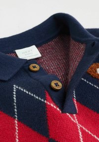 Polo shirt bleu marine avec un motif en losanges rouges et blancs, featuring des boutons en bois et une texture tricotée douce. L'étiquette indique "NEXT baby."