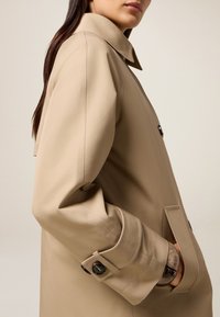 Beige trenchcoat met een gestructureerd ontwerp, grote kraag en knoopmanchetten. Voorzien van een zijzak en een gladde, matte textuur.