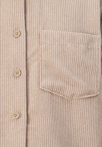 Tessuto in corduroy beige con righe verticali, dotato di tasca sul petto e tre bottoni beige sul davanti. Texture liscia, design casual.