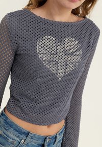 Grå, langermet, kort jakke laget av åpenstrikket stoff med et hjerte-design i midten. Uformell stil med ribbestrikkede kanter.