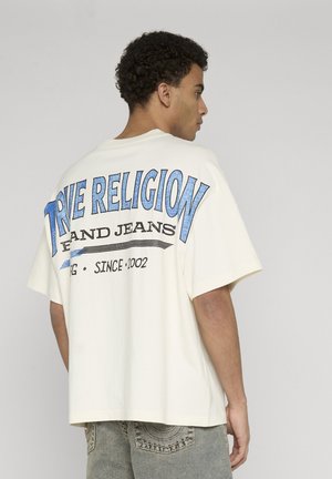 Młody mężczyzna o kręconych włosach, ubrany w białą, oversizową koszulkę z niebieskim napisem "True Religion" oraz jasne dżinsy, stojący bokiem.