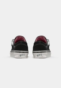 Zapatos de lona negra de corte bajo con forro rosa, suelas de goma blanca y costuras blancas en contraste. Presentan un acento metálico circular en el talón.