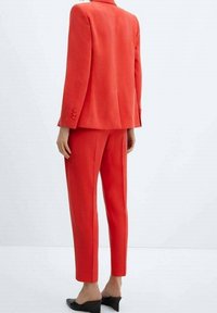 Costume rouge ajusté avec un blazer à un bouton et un pantalon fuselé. Tissu lisse, minimalisme dans les détails, accompagné de chaussures noires à enfiler.