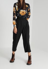 Schwarze, lockere, cropped Hosen kombiniert mit einer durchsichtigen schwarzen Bluse mit floralen Mustern in Orange und Gelb. Braune Schnürstiefel.