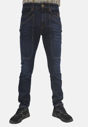 Jeans in denim blu scuro con cinque tasche, chiusura con bottone e cuciture visibili. Indossati con scarpe da ginnastica nere.