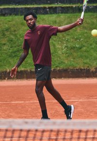 Tennis pemain mengenakan kaos merah maroon dan celana pendek hitam, dengan raket tenis di tangan, di lapangan tanah liat dengan latar belakang hijau.