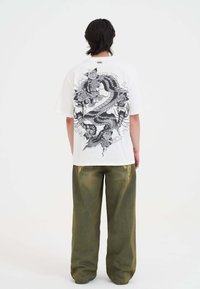 Witte oversized t-shirt met zwarte draak graphic op de achterkant, gecombineerd met losse, groene textuur broek met subtiele vervaagdetails.
