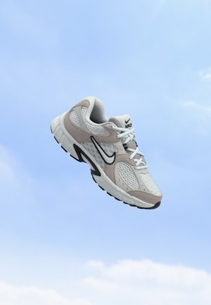 Chaussure de course Nike blanche et beige avec des accents noirs et des lacets flottant sur un fond de ciel bleu clair.
