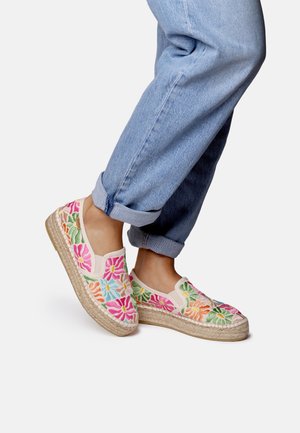 Fødder iført slip-on espadriller med farverig blomsterbroderi, kombineret med opkørte lyseblå jeans mod en ensfarvet baggrund.