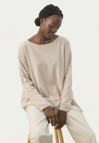 Beiger, übergroßer Pullover mit weitem Ausschnitt, langen Ärmeln und gerippten Abschlüssen, kombiniert mit hellen Hosen. Weiche Textur, lockere Passform.