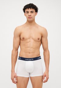 Boxer briefs brancos para homens com uma cintura azul-marinho que apresenta o logo "LACOSTE". O design é suave, ajustado e elástico para maior conforto.