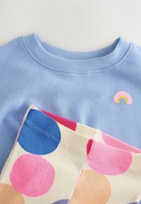 Ljusblå sweatshirt med ribbad rundhals och ett litet regnbågsapplikation. Nedan, mönstrade leggings med stora rosa, blå och orange prickar.