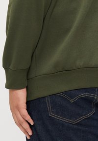 Sudadera verde oliva con puños y dobladillo acanalados, combinada con jeans azul oscuro que presentan costuras amarillas y un diseño de bolsillo trasero en forma de corazón.