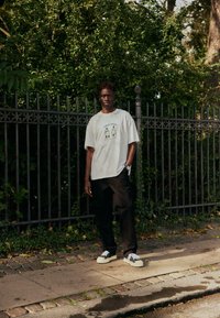 T-shirt blanc surdimensionné avec motif, pantalon noir, baskets blanches et noires, debout sur un trottoir près d'un décor feuillu vert et d'une clôture noire.