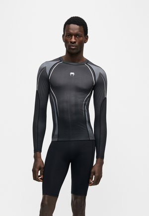 Athletischer Mann, der ein eng anliegendes langärmeliges Sportoberteil in Schwarz und Dunkelgrau sowie schwarze knielange Kompressionsshorts vor einem schlichten Hintergrund trägt.