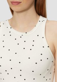 Bílý top na ramínka s černými puntíky, žebrovaná struktura, kulatý výstřih a široké ramínka, ukazující přiléhavý design bez dalších detailů.