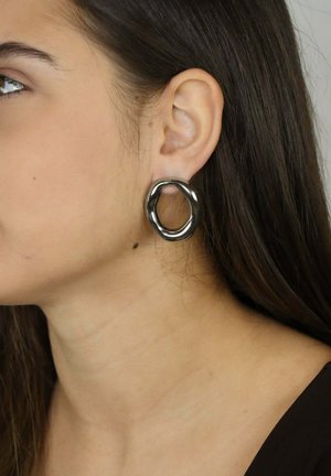 Grande orecchino a cerchio testurizzato in colore metallico scuro, con un design irregolare che aderisce strettamente al lobo dell'orecchio.