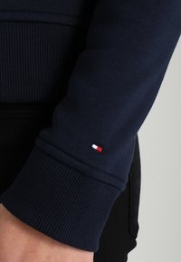 Sudadera azul marino con puños acanalados. Presenta un pequeño logo bordado en rojo, blanco y azul en la manga. Textura de tela suave.