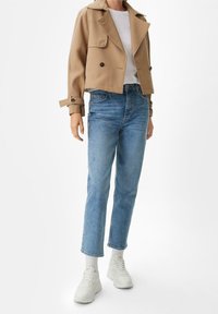 Chaqueta corta beige con botones de doble pecho, camiseta blanca debajo, combinada con jeans rectos de mezclilla azul claro y zapatillas blancas.