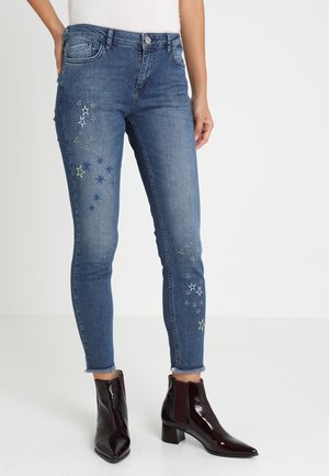 Smaltsiddende blå jeans med broderede stjernemønstre på lårene og frynset kant, båret med skinnende mørkebrune ankelstøvler.