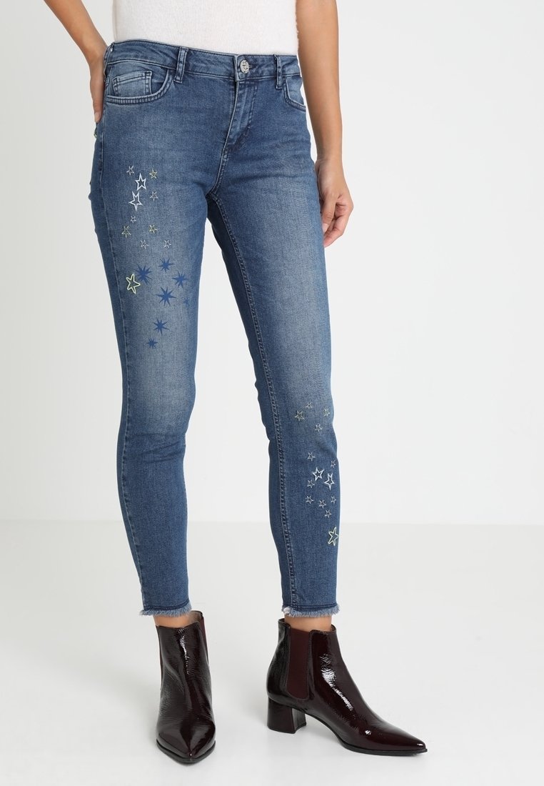 Jean slim bleu avec des motifs d'étoiles brodés sur les cuisses et des ourlets effilochés, porté avec des bottines marron foncé brillantes.