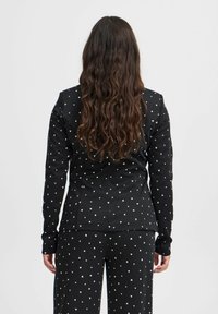 Kvinna med långt, vågigt brunt hår som bär en svart kostym med små vita hjärtmönster, vänd bortåt mot en enkel ljus bakgrund.