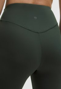 Magasabban álló, sötétzöld, aktív leggings, sima, rugalmas anyagból készült, lapos derékkal és diszkrét logó részletekkel.