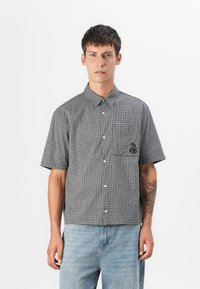 Short-sleeve checkered shirt in black and white, featuring a button-down collar, front pocket, and embroidered design, paired with light denim jeans.